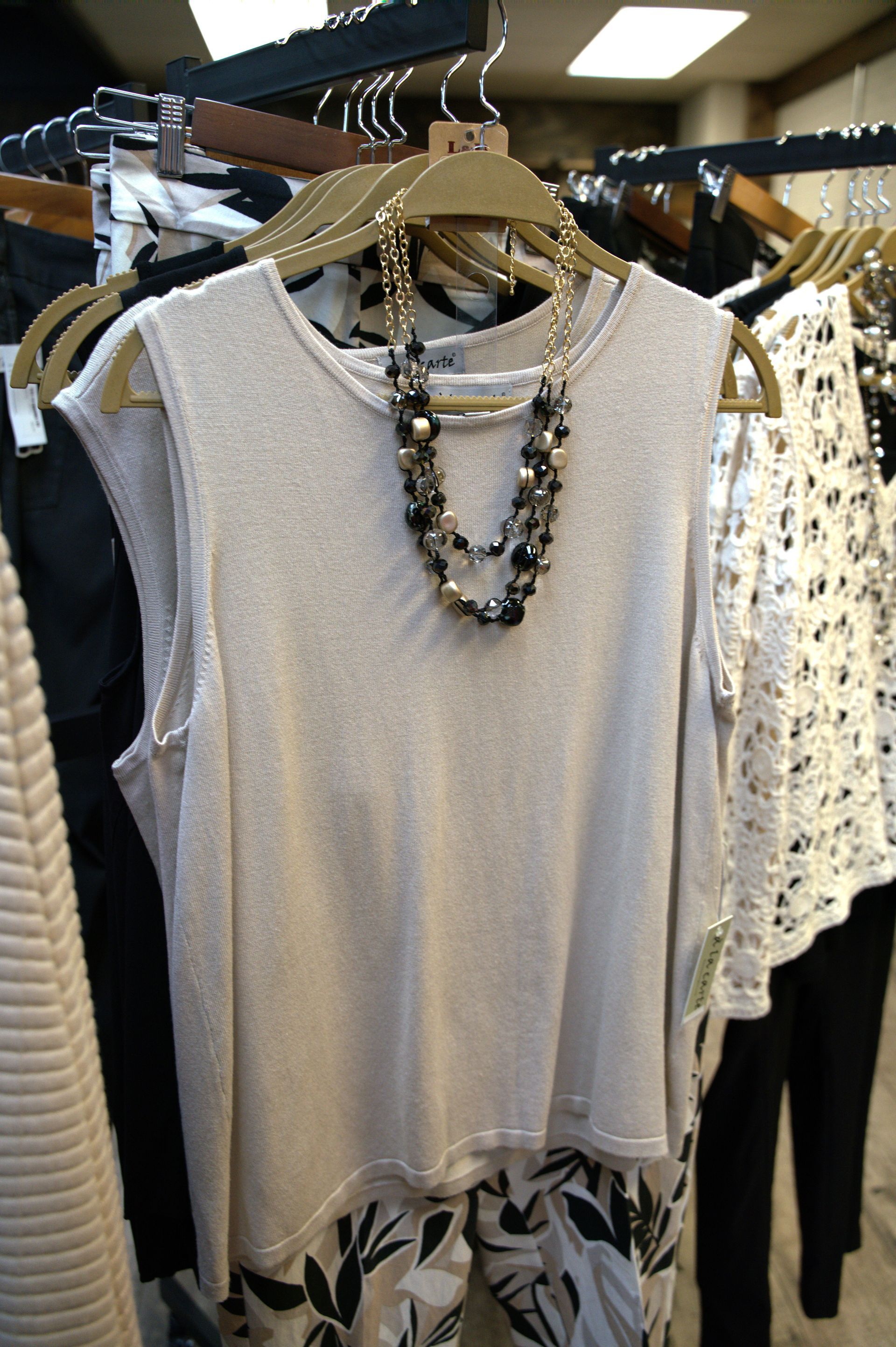 Beige sleeveless top with beaded necklace hanging on a clothing rack, other garments in the background.