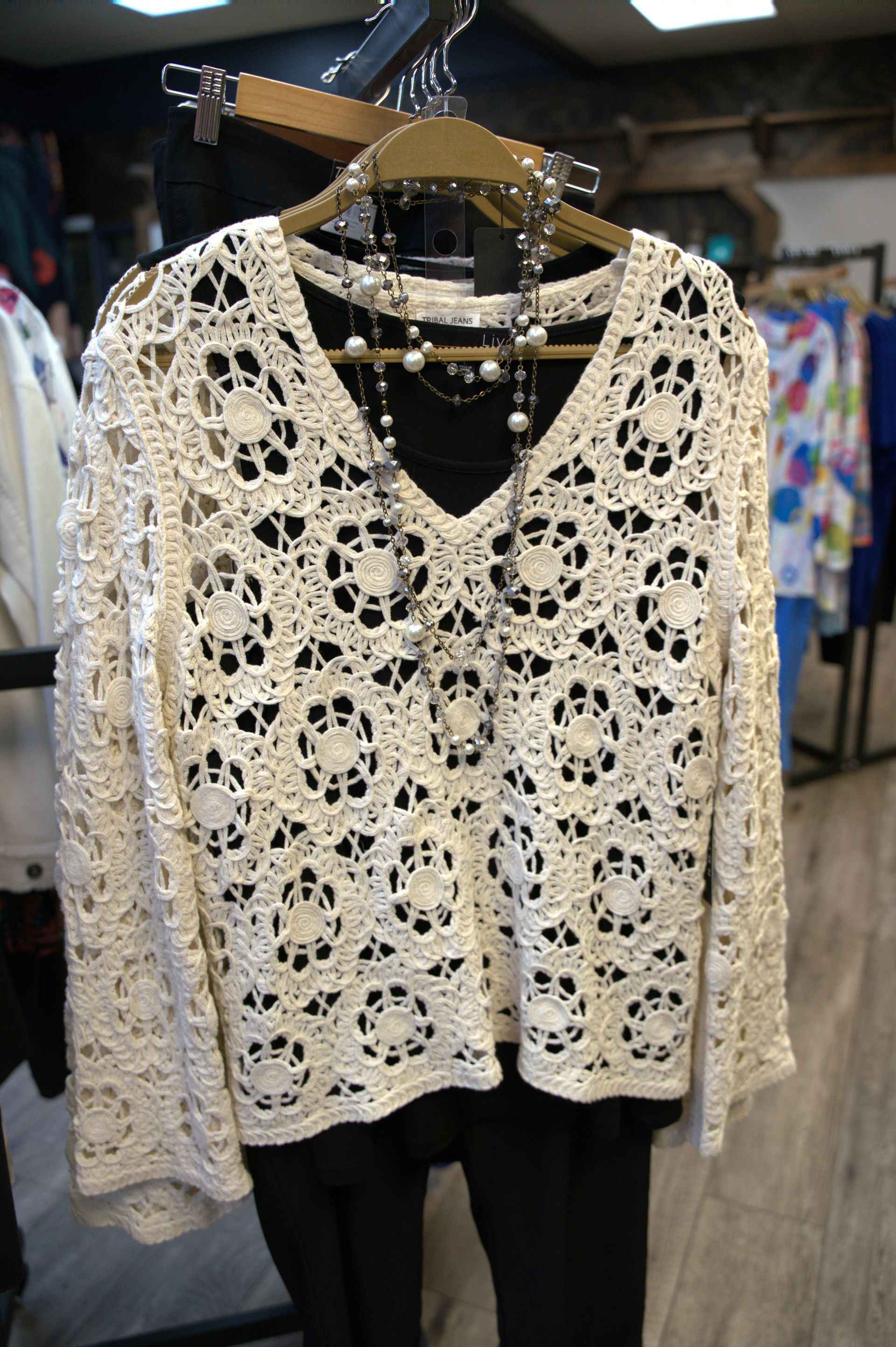 White crocheted sweater with floral patterns, on a hanger with a black shirt and pants underneath.