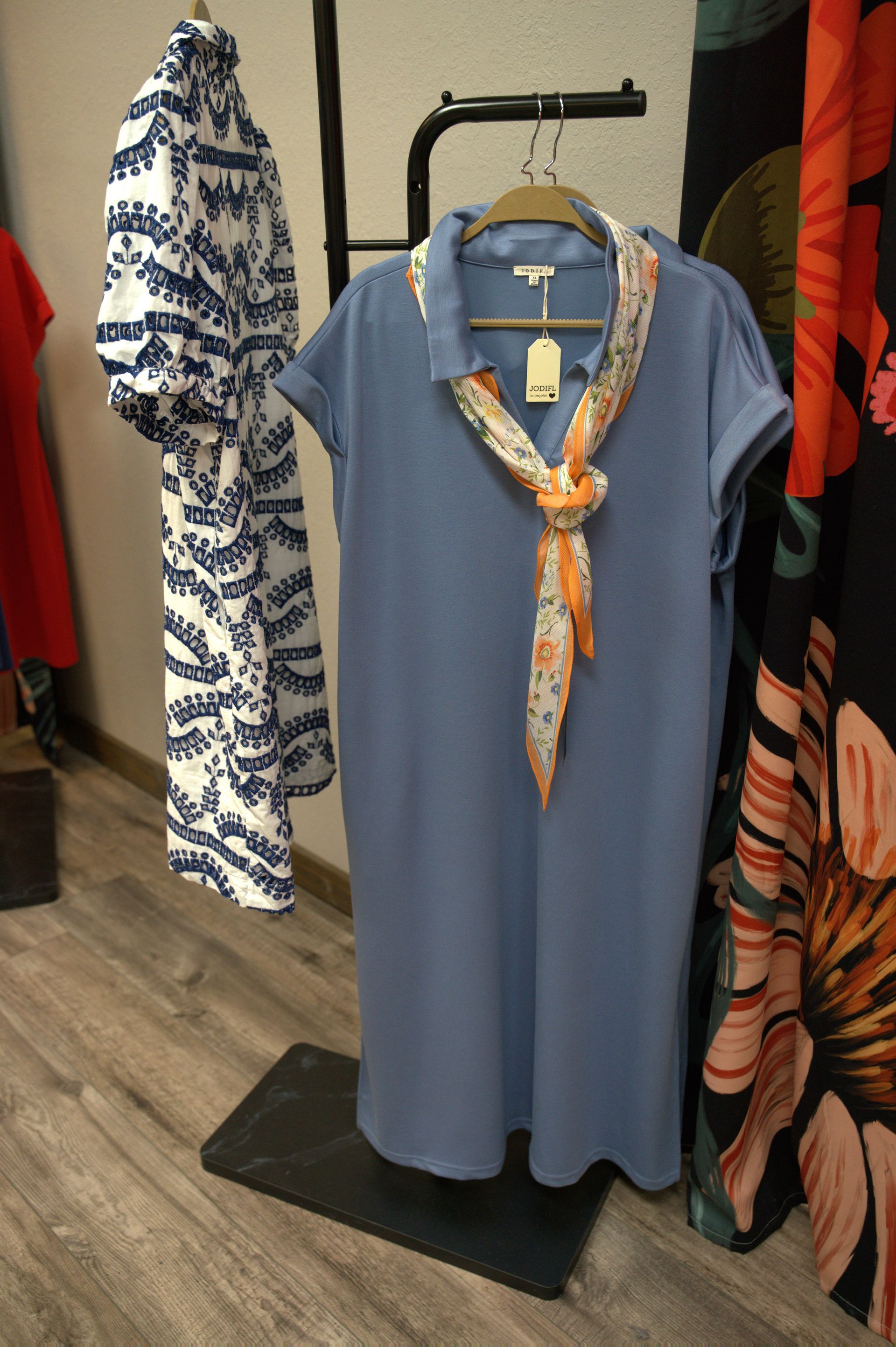 Clothes rack displaying a blue dress with an orange scarf. A white dress with blue print hangs to the left.
