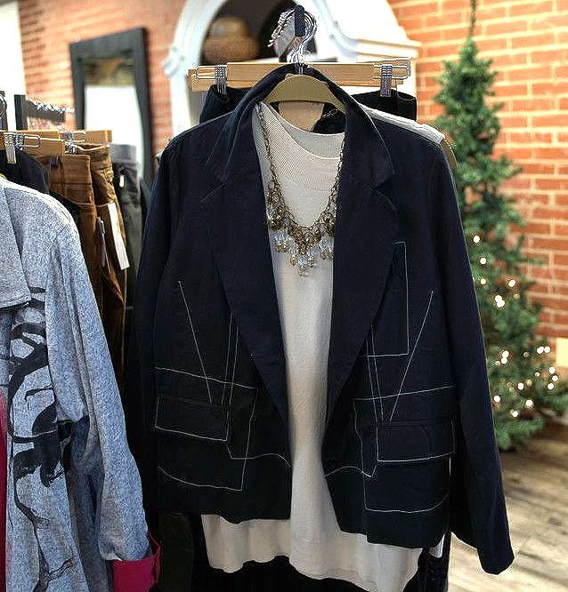 A navy blazer with white stitching and a white blouse on a clothing rack inside a store with a brick wall.