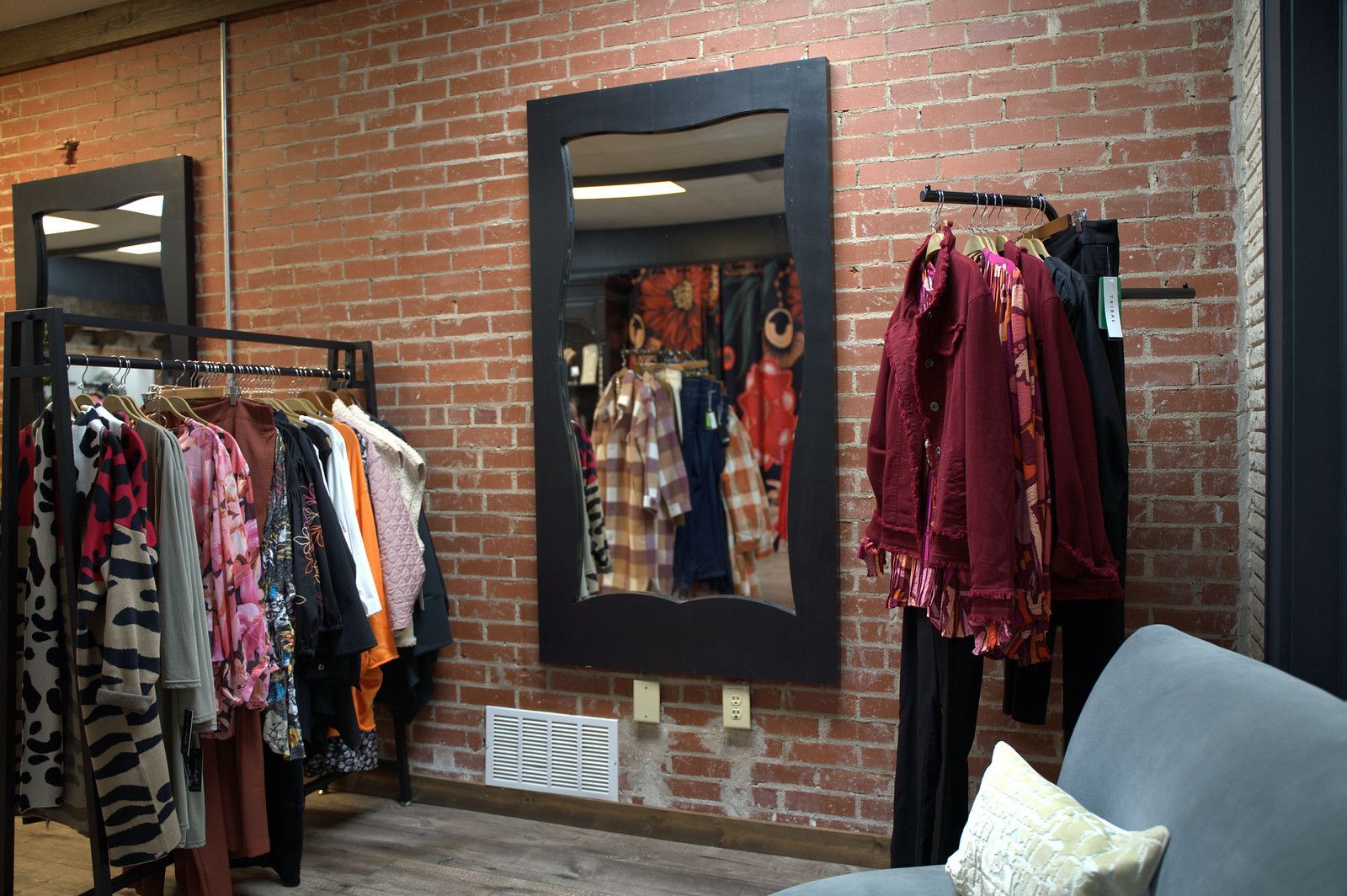 Clothing racks and a mirror in a boutique with exposed brick walls. The mirror reflects more clothing and a patterned shirt.