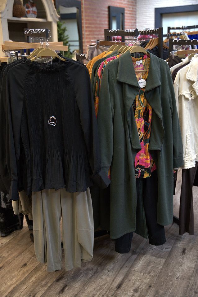 Clothing racks in a boutique. Black blouse and tan skirt on left, green coat over patterned top on right. Wooden floor.