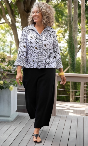 Woman with curly gray hair wearing a black and white floral blouse and black pants, standing on a wooden deck outdoors.