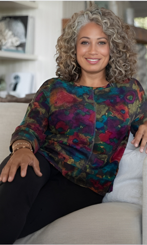 Woman with curly gray hair smiles, wearing a colorful floral top and black pants, seated on a couch indoors.
