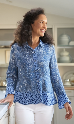 Woman in a blue patterned shirt and white pants, smiling. Leaning on a counter in a well-lit kitchen.