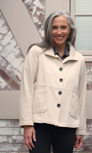 Woman with gray hair in a beige jacket smiling in front of a brick wall and window.