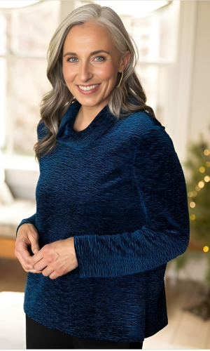 Woman with silver hair wearing a textured blue top, smiling, indoors with a Christmas tree visible.