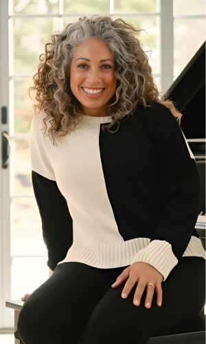 Woman with curly gray hair smiles, wearing a black and cream sweater, sitting near a piano.