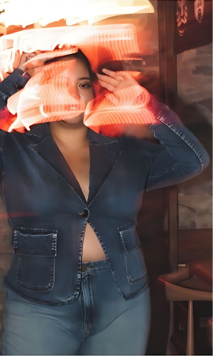 Woman in denim jacket, hands near face, partially obscured by blurred orange light; standing near a wooden wall.