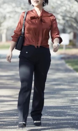 Woman in rust-colored blouse and black pants walks on a pathway; she carries a black purse.