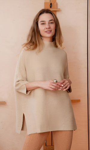 Woman in a beige sweater and pants smiles at the camera. She stands in a light-colored room.