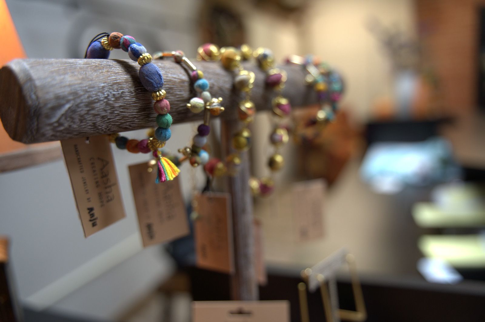 Bracelets on a wooden display stand. Colorful beads and gold accents, tags attached. Blurry shop interior in the background.