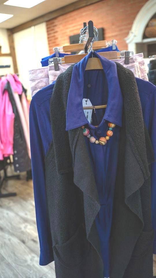 Clothing rack with a blue top, dark vest, and beaded necklace. The store interior features brick and wooden accents.