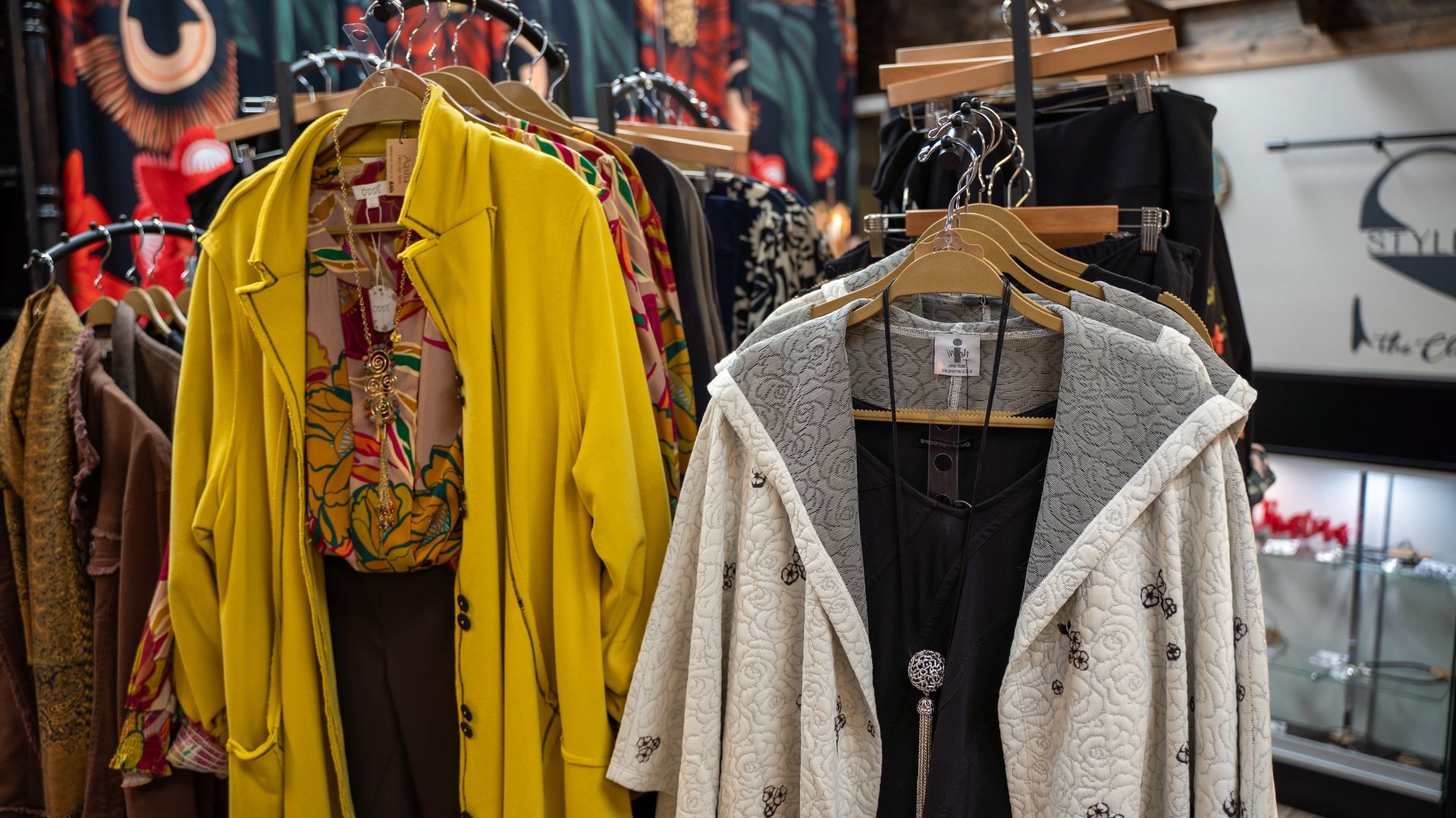 Clothing rack in a shop displays a yellow coat and a white hooded cardigan, along with other colorful garments.