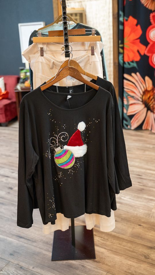 Black top with Christmas ornament and Santa hat design on a clothing rack.