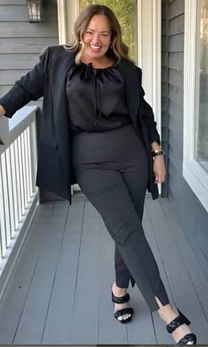 Woman smiles, wearing black blazer, top, and pants, standing on a porch. She crosses her legs, wearing braided sandals.
