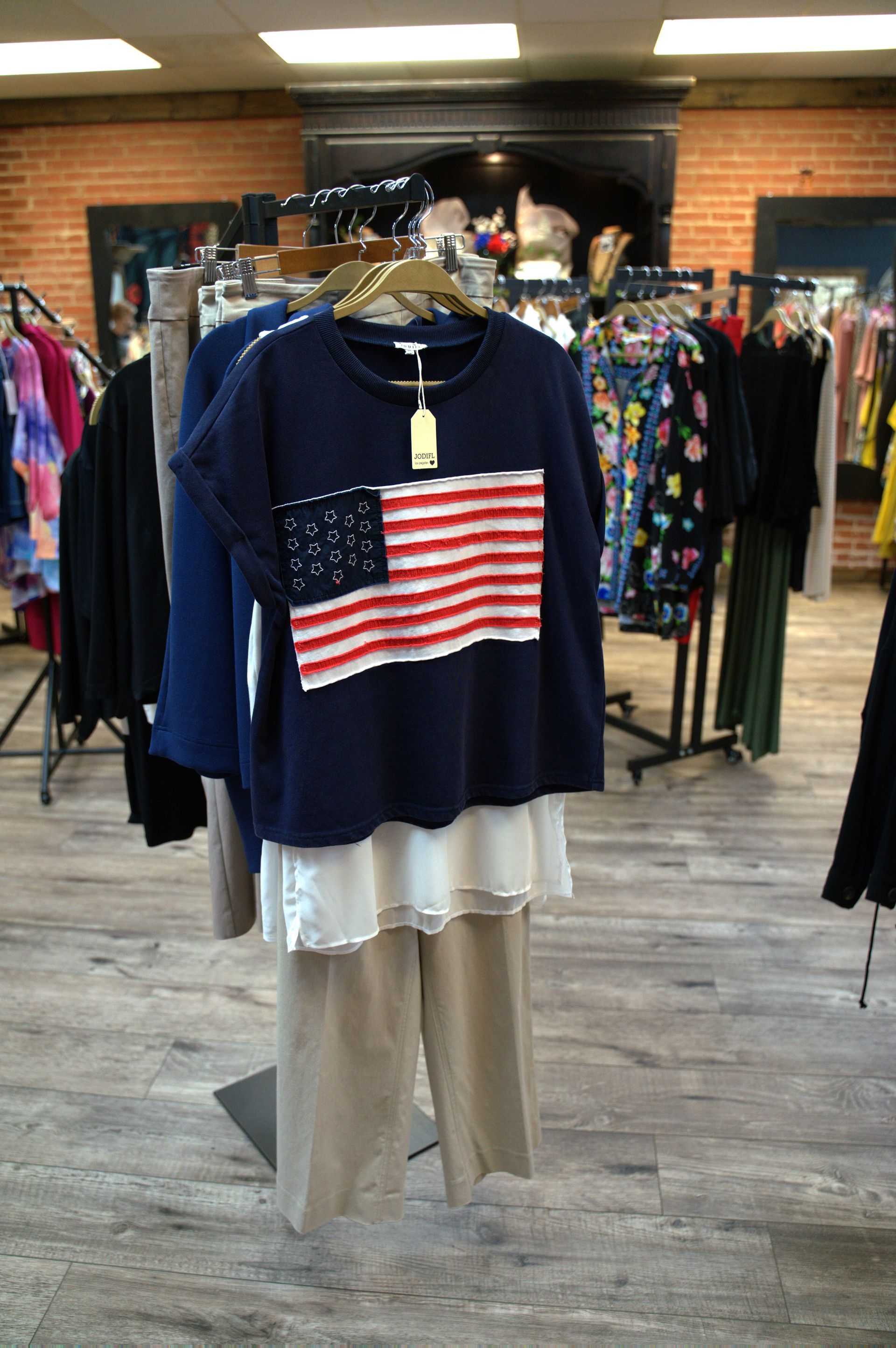 Outfit display: Navy blue top with American flag design, white layered top, tan pants on a rack in a boutique.