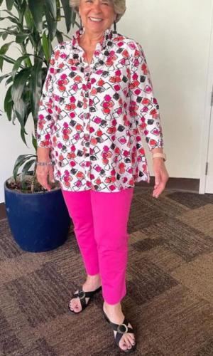 Woman in a floral print top and pink pants, wearing black sandals, standing indoors near a plant.
