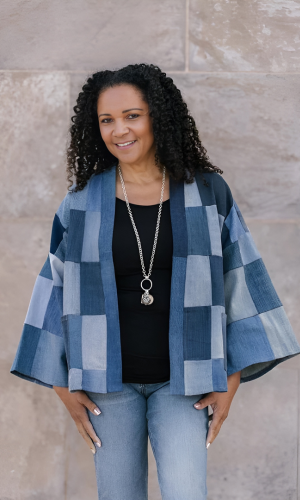 Woman with curly hair wearing a patchwork denim jacket, black top, and jeans, standing in front of a stone wall.