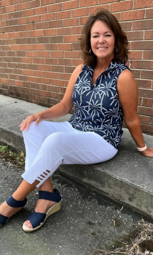 Woman in white pants and a palm-tree-print navy top sits smiling against a brick wall. She wears navy sandals.