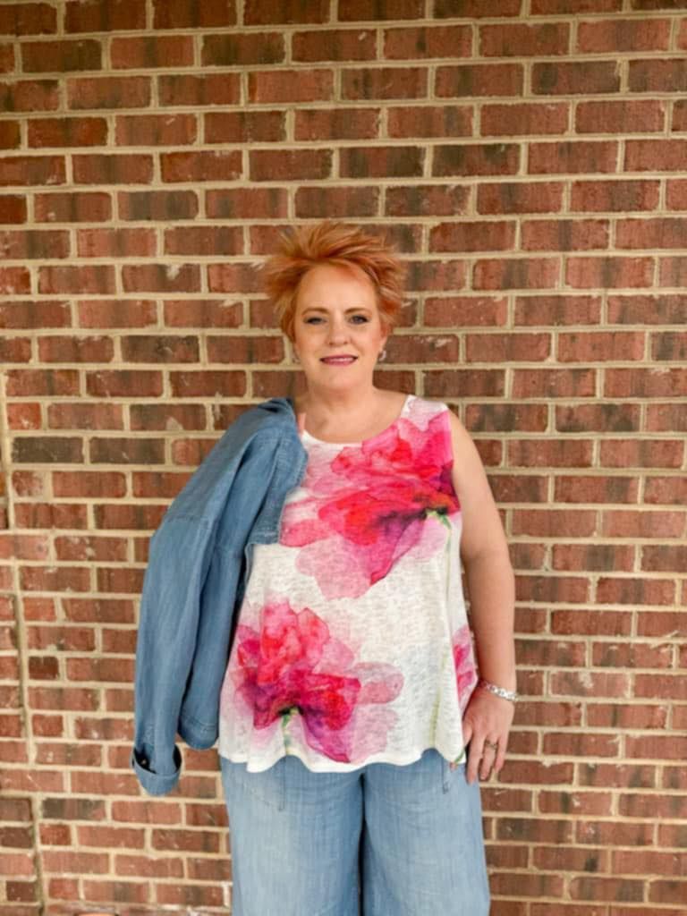 Woman with short red hair wearing a floral top and jeans stands in front of a brick wall, holding a blue jacket.