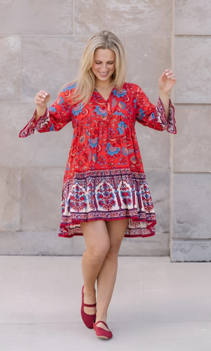 Blonde woman smiles, wearing a red floral dress and red flats, outside a building.