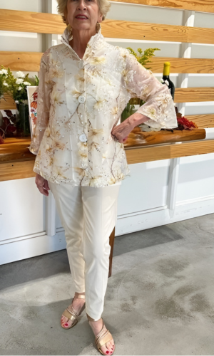 Woman in cream-colored outfit, including sheer floral top, poses indoors. She wears sandals, has short hair, and smiles.