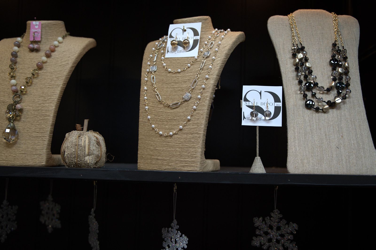 Jewelry display on a black shelf with necklaces and earrings on burlap bust forms; snowflake decorations below.