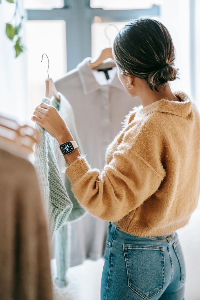 Woman looking at clothes on hangers, wearing a yellow sweater and jeans, near a window.