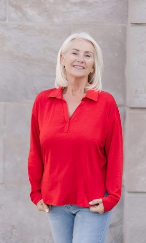 Woman with blonde hair wearing a red top and blue jeans smiles outdoors against a stone wall.