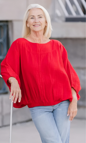 Woman with blonde hair smiles, wearing a red blouse and jeans, leaning on a white railing outdoors.