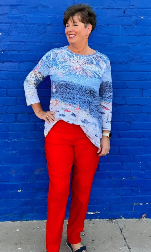 Woman in red pants and a blue patterned shirt poses in front of a blue wall, smiling.