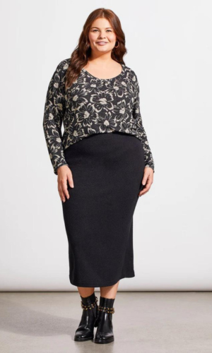 Woman in plus-size floral top and black skirt, standing in a studio. She wears boots and smiles.