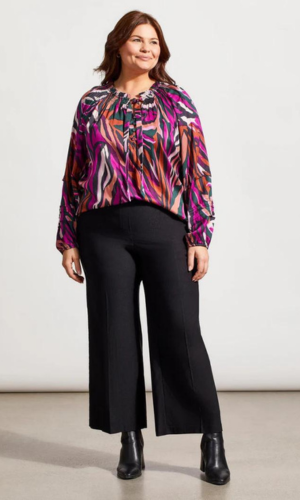 Woman in a colorful patterned blouse and black culottes, standing in a studio, smiling.