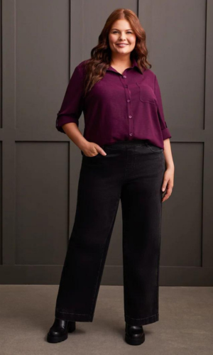 Woman in a purple shirt and black wide-leg jeans, smiling. She is standing in front of a gray wall.
