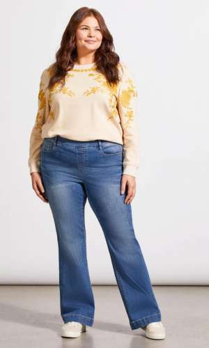 Woman in cream sweater with yellow embroidery and blue jeans, smiling, standing against a white background.