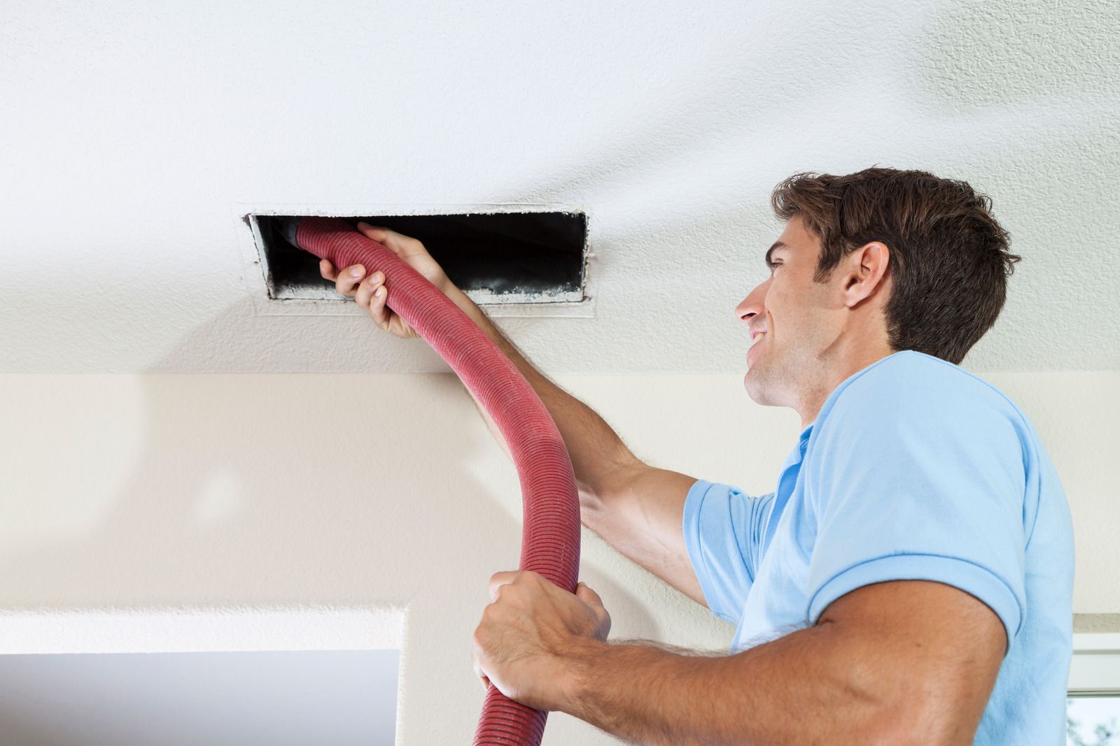 Man cleaning a ceiling vent with a hose, inside a home.