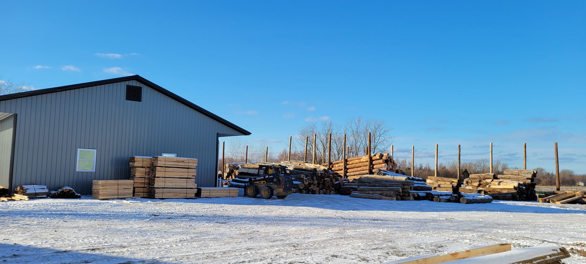 A snowy lumberyard with a gray building, stacked lumber, and logs under a clear blue sky.