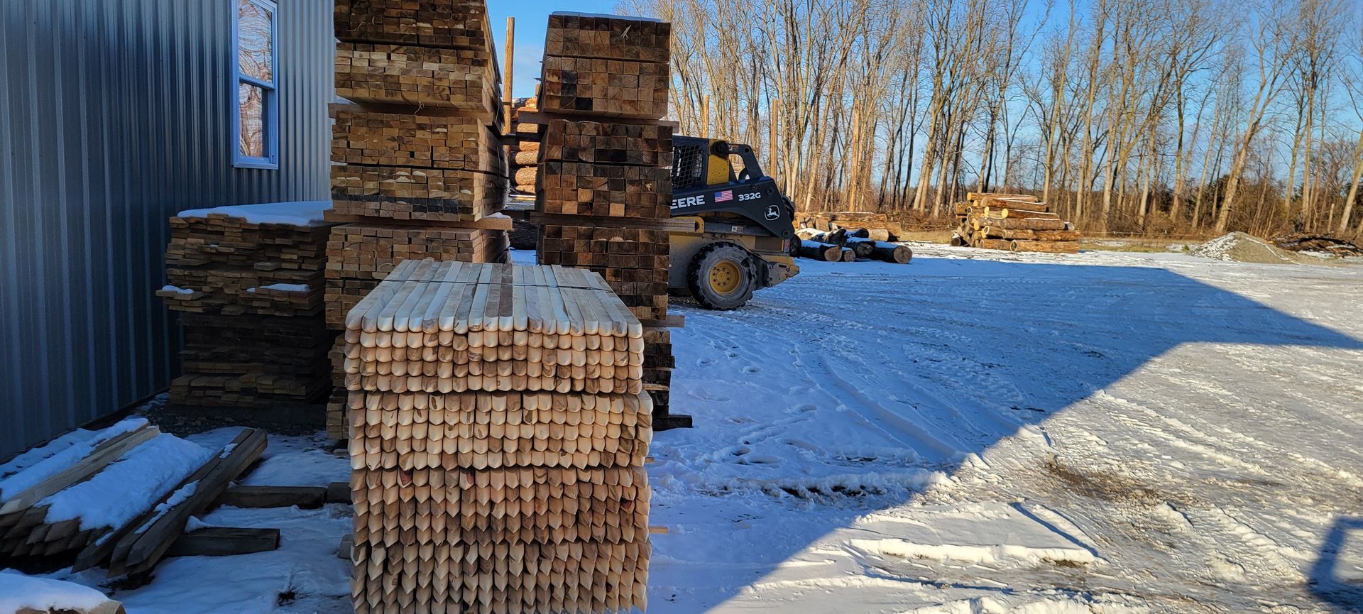 Lumberyard scene: stacks of wood, small front loader, snow-covered ground, building, bare trees, and a clear blue sky.