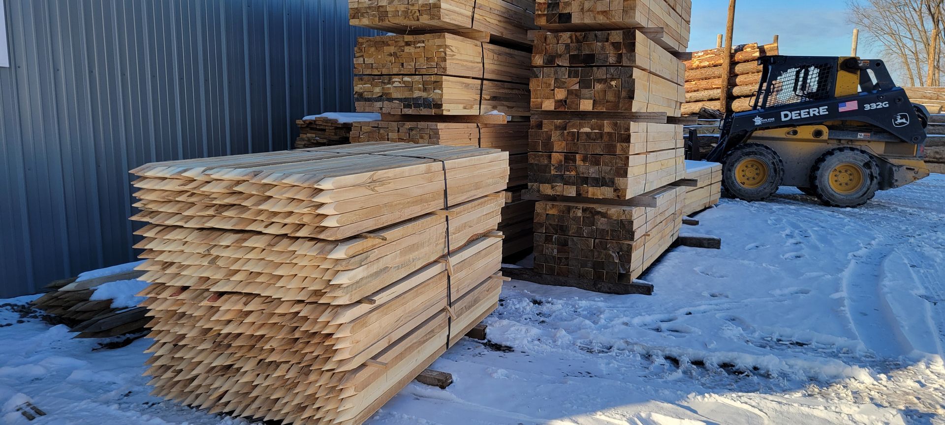 Stacks of lumber in snowy outdoor setting with a small skid steer loader nearby.