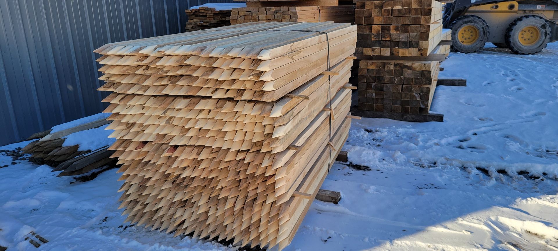 A large stack of wooden stakes sits on snow-covered ground near a building and a piece of heavy machinery.