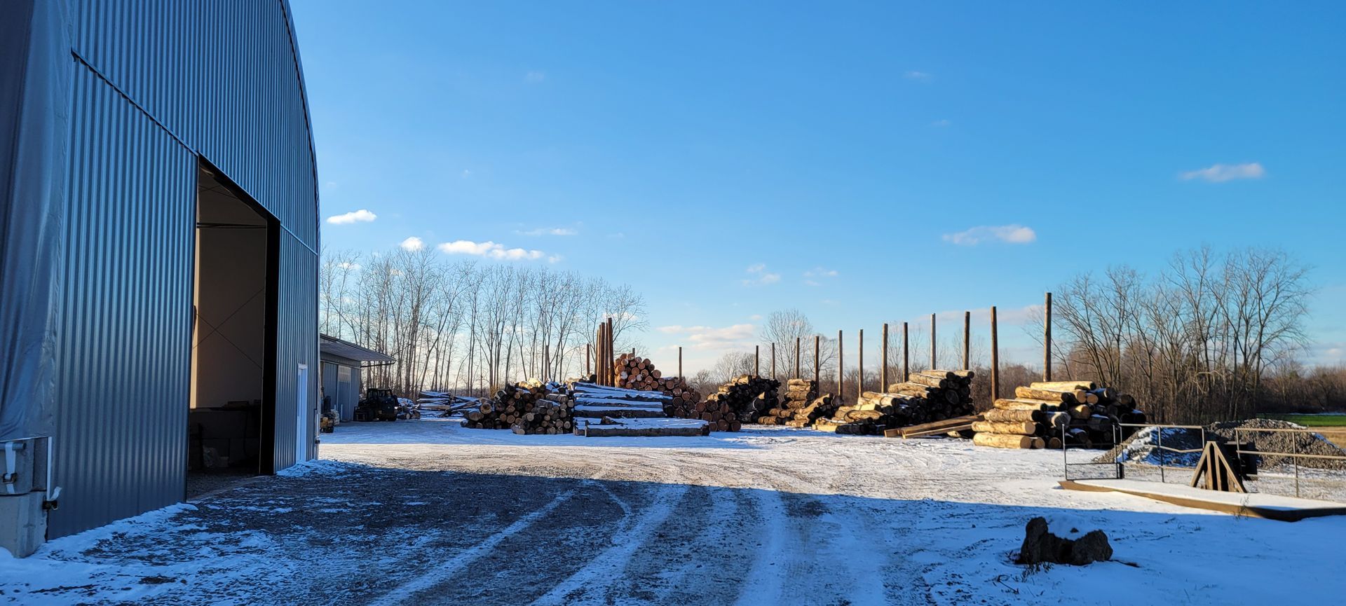 Snow-covered yard with a large metal building on the left. Lumber piles and trees are visible under a clear blue sky.