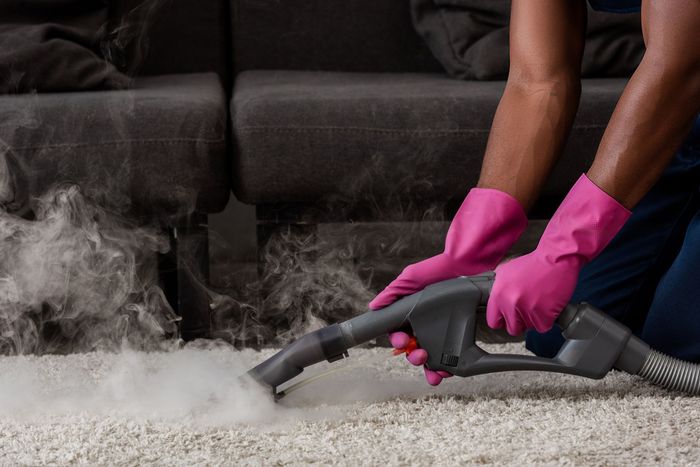 Person with pink gloves vacuuming a dusty carpet beside a couch