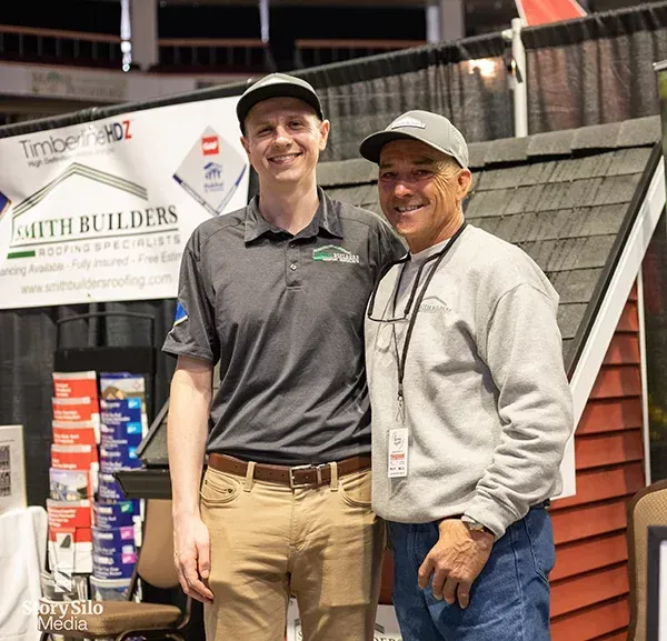 Two men at a trade show booth with a roof display, one wearing a cap and smiling at the camera.