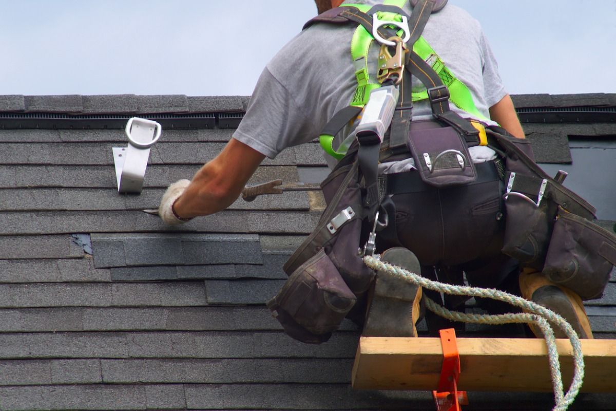Roofer wearing safety harness repairs shingles on a roof; holding a hammer and tool belt.
