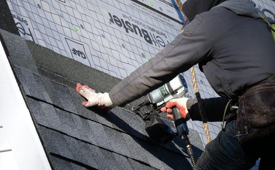 Roofer using a nail gun to attach shingles to a house roof, wearing gloves and a safety harness.