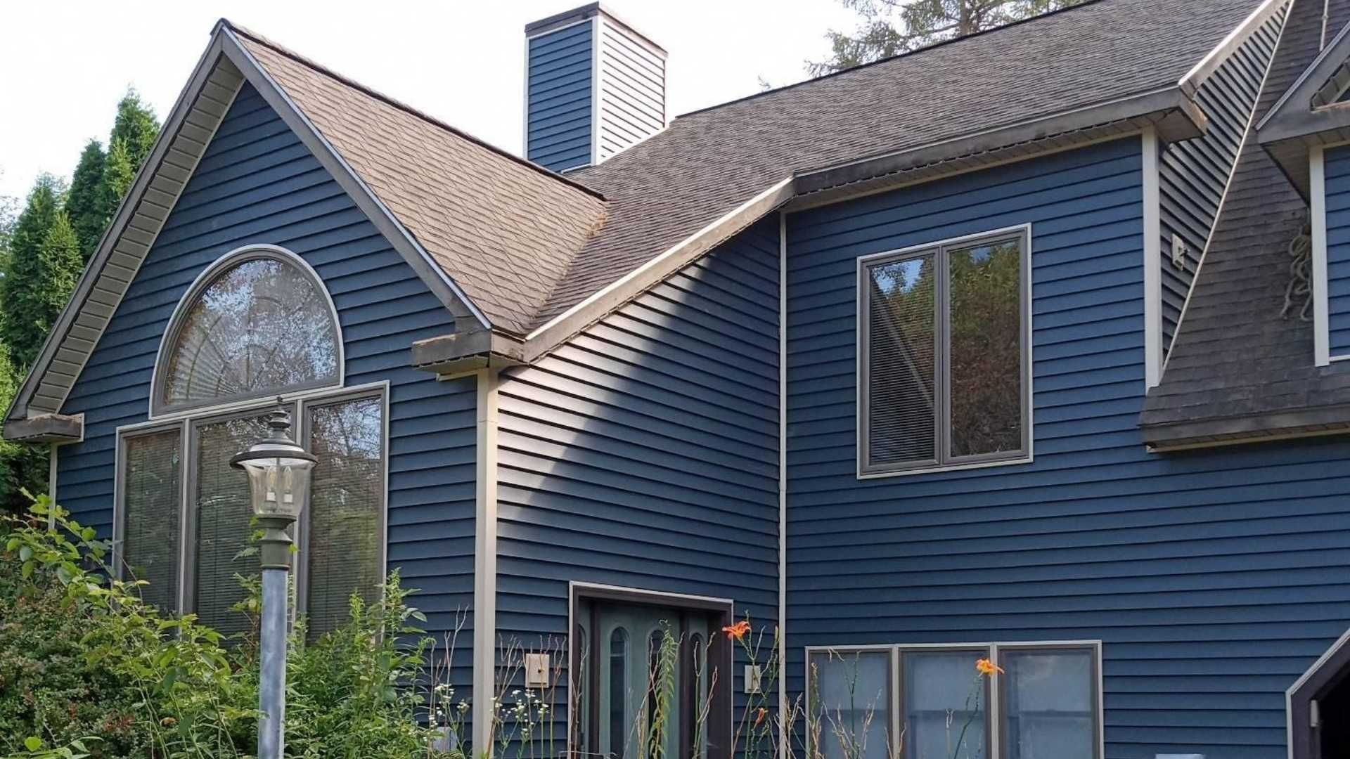 Blue sided house with brown roof, multiple windows, and a chimney.
