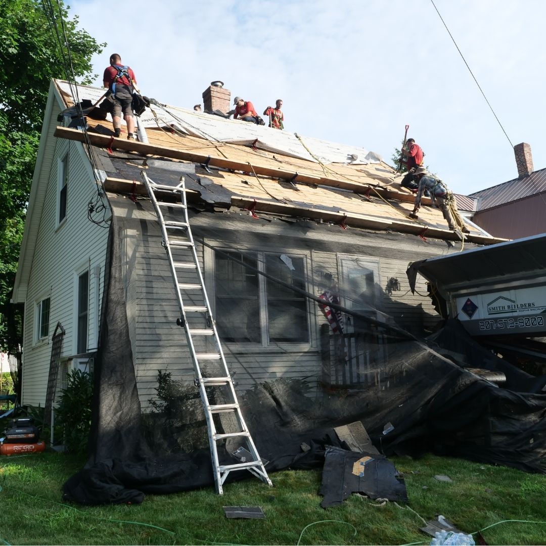 Roofers in red shirts remove shingles from a white house roof; a ladder leans against the side.