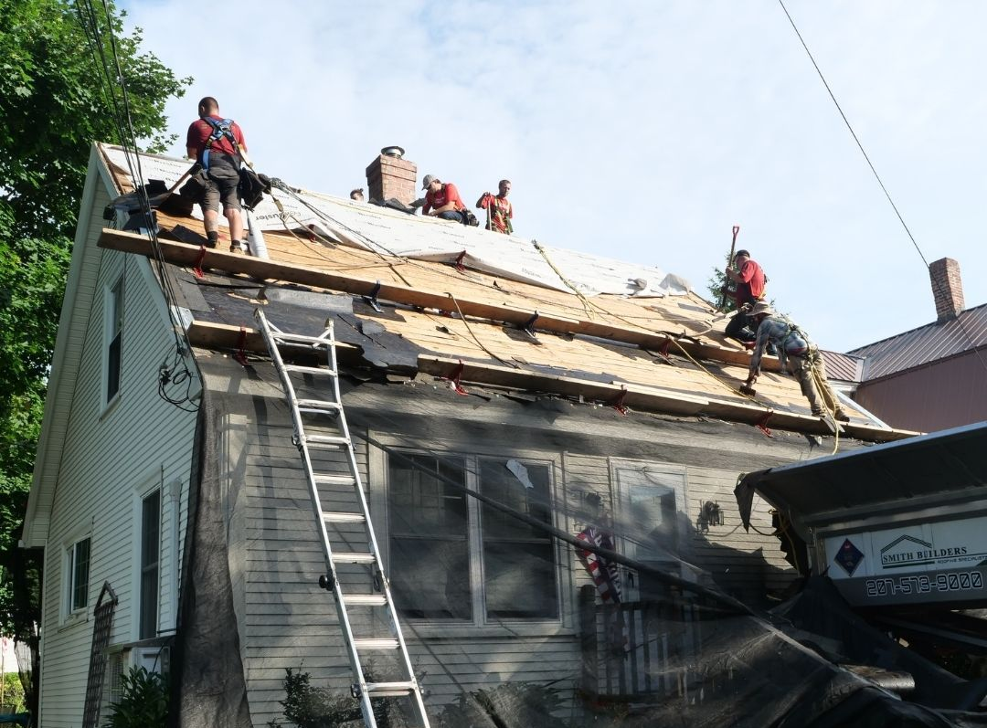 Roofers working on a house with scaffolding. They wear safety harnesses and work on a partly-demolished roof.