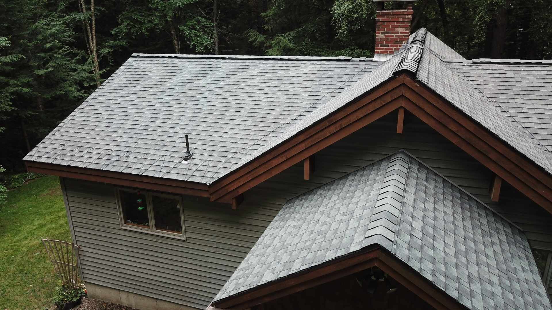 A roof with gray shingles, brown trim, and a brick chimney on a house, viewed from above in a forest.